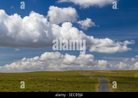 Narrow winding road crossing open moorland beneath a dramatic blue sky filled with billowing cumulus clouds Stock Photo