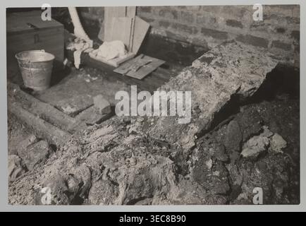 Vilnius. Cathedral — interior of the royal crypt. Photograph by Jan Bułhak 1876–1950, 1931. Depicts crypts revealed by archaeological excavations after the 1931 flood in Vilnius. Photograph from nature; 14.5 × 16.5 cm., A grayscale photograph showing a rough indoor space with a brick wall backdrop and a dirt floor scattered with broken concrete and rubble; a large irregular slab of concrete or stone leans across the right side, and a pile of crumbled material with coarse texture occupies the foreground. Toward the left a metal bucket sits near a wooden chest and a group of wooden boards and Stock Photo