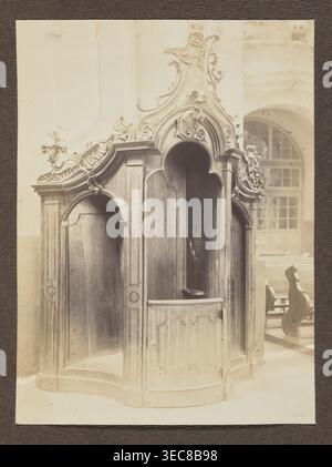 Confessional in the Dominican Church; photograph, Bułhak, Jan 1876–1950; Dominican Church of the Holy Spirit Vilnius; confessionals; Baroque; ca. 1914; photograph, photographic print: height 16.0 cm, width 11.9 cm; album page: height 31.0 cm, width 23.5 cm, The image shows an ornate wooden confessional booth with carved decorative scrollwork and curved arches, finished in warm brown tones; the interior appears darker brown and shadowed. The booth sits on a pale floor and is set against a background wall and large window area rendered in light beige and cream tones. In the background to the Stock Photo
