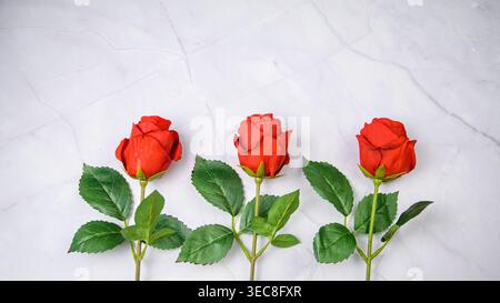 Three red roses with green leaves on light marble, clean romantic composition with upper space for design or text. Stock Photo