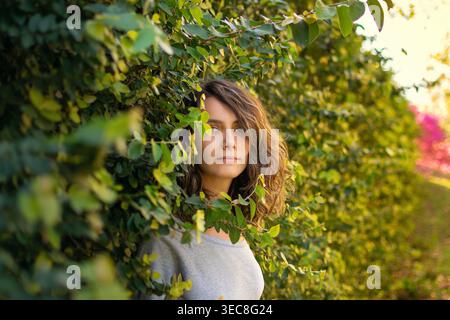 A young woman smiles while standing in front of a green wall covered with various plants and leaves. Bright sunlight shines through the foliage, creat Stock Photo