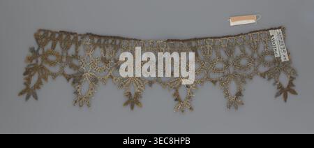 Lace, bobbin lace, 16th–17th century, Flanders, fabric, width 7 cm, length 27 cm;, A delicate lacy trim with an openwork pattern lies flat against a gray background, showing an ornate repeating design of loops, floral shapes, and dangling leaf-like motifs; the lace appears in shades of metallic gold and darker brown, with subtle variations creating a textured, slightly aged look, and a small fabric tag in beige and white is attached near one end. Stock Photo