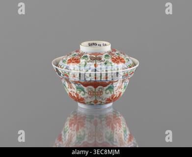 Bowl with a lid; porcelain, colored; decorated with bats, longevity shou, lotus tendril ornament, lotuses, and peaches; Qing dynasty, Qianlong period/style, 1736–1795; China; height 9 cm, diameter 10.7 cm; maker: Ignacy Jan Paderewski (1860–1941)., A small lidded porcelain bowl with a rounded body and narrow foot sits centered against a smooth gray background, its glossy surface reflecting beneath it; the bowl and lid are decorated all over with intricate floral and foliate patterns in colors including red, green, blue, and a touch of yellow on a white base, with repeating motifs and stylized Stock Photo