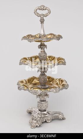 Etażera, Fraget, Józef (Warsaw; silver and plated goods factory; 1824–1945), tableware elements. Etażera — three-tiered platter on a three-sided base, with three knobbed plates, plated with gold on the outside and silver on the bottom, with silver-plated tin overlays on the edges. Finished with an oval decorative handle. Composed of two parts: the base and a plate holder inserted into a cylindrical opening. Marked with an oval Fraget mark with the date 1843. Vessel, 1843, Poland, Warsaw; height 57 cm, width 25.9 cm., A three-tiered ornate metal stand with a decorative handle at the top, Stock Photo
