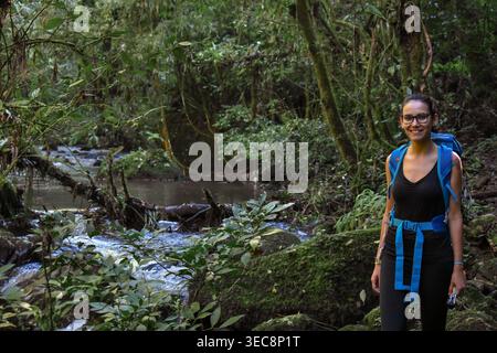 A young woman looks at the camera while standing next to a stream in the jungle. She holds an action cam in one hand and wears a backpack. She is read Stock Photo