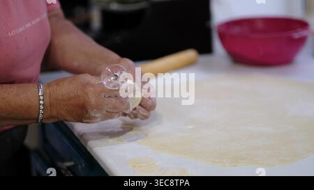 Glass rim cuts multiple round shapes from rolled dough as hands prepare pastry on a kitchen counter. The scene illustrates homemade cooking activity Stock Photo