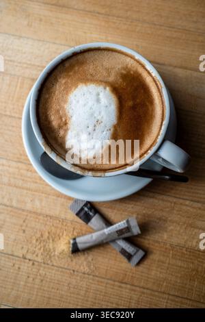 An overhead view of a coffee flat white and two paper sachets of demerera suger on a table in a cafe in England in the UK. Stock Photo