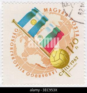 HUNGARY - 1962 May 21: 1.70 forint ochre and bistre postage stamp commemorates the 1962 FIFA World Cup hosted in Chile, specifically highlighting the national flags of Argentina and Bulgaria. These teams competed in the same group during the opening stage of the tournament. The background features an orange-tinted globe focused on the Western Hemisphere to denote the South American venue. A classic yellow leather football is positioned in the foreground. In this specific tournament, Argentina failed to progress past the group stage, while Bulgaria was also eliminated early Stock Photo