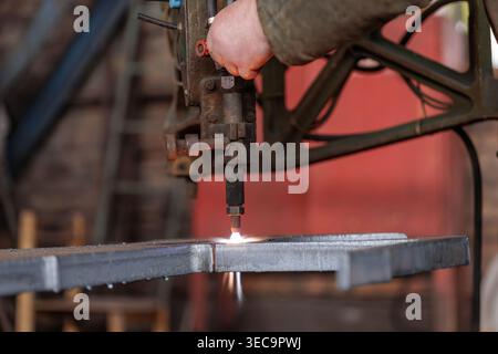 Worker operating plasma cutting torch on metal plate in industrial workshop. Manufacturing process with sparks and heat in heavy industry. Stock Photo