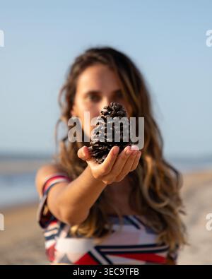 A woman stands on the sand by the water. She holds a pine cone in her hand. The sun shines bright, and the beach stretches behind her. The scene shows Stock Photo