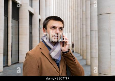 Man dressed in stylish coat with scarf talking on phone while walking through modern city architecture in business setting. businessman, manager, lawy Stock Photo