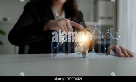 Business woman interacts with a glowing light bulb on a desk, symbolizing ideas while analyzing digital graphs and data sets, representing innovation Stock Photo
