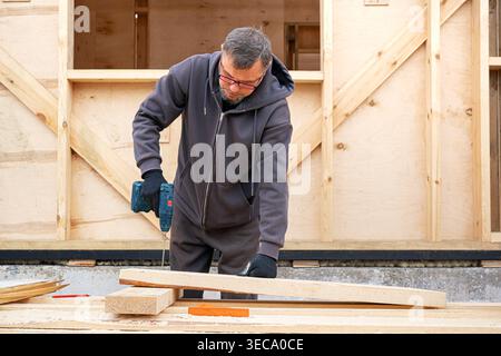 Carpenter man using electric screwdriver to fasten wooden planks. Work in progress near a frame house under construction. DIY eco wooden home concept. Stock Photo