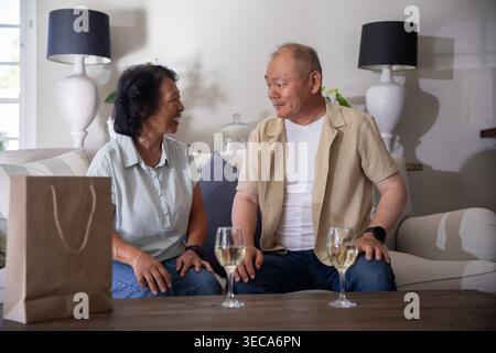 Senior Asian woman, mature Asian man sitting on sofa at home smiling, conversing with wine glasses Stock Photo