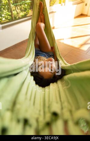 A young woman lies in a green hammock, looking at the camera with a relaxed expression. She is indoors, surrounded by natural light and greenery. Stock Photo