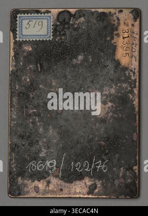 Title: IESVS MATRIS DELICIAE IESVS PATRIS SOLATIVM (The Heavenly and Earthly Trinity), A rectangular metal plate with rounded corners rests on a neutral gray background; the plate shows a worn, mottled surface with large areas of dark gray and black corrosion interspersed with exposed coppery brown patches and a few pale pinkish spots, and there is a decorative blue-and-white rectangular label affixed near the top left corner; along the top right edge the metal is cleaner and more copper-colored, and the plate surface has a smeared, scratched texture with several small pits and dark blotches Stock Photo