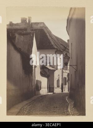 Bernardine Alley; photograph, Jan Bułhak (1876–1950), 1912, Vilnius; photographic print: 15.7 × 11.5 cm; album page: 31.0 × 23.5 cm., A narrow cobblestone street flanked by tall buildings with textured facades, showing a group of rooflines and chimneys in the background; the left building has a dark vertical patch of vegetation or shadow, and the right side features a mounted lantern and other wall-mounted fixtures. The scene displays muted tones of beige, brown, cream, and sepia, with soft tonal gradients and areas of light and shadow along the street and building surfaces. Stock Photo