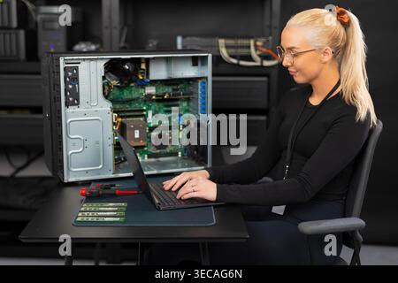 Female IT technician in her twenties works on laptop beside open server for diagnostics Stock Photo