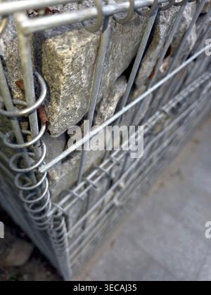 Construction workers build a gabion wall using mesh and stones at a project site during daylight. The wall serves as a retaining structure for stabili Stock Photo