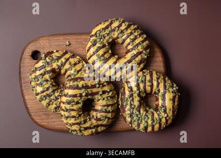 Three delicious donuts topped with pistachios and drizzled with chocolate sit on a wooden board against a textured background. Stock Photo