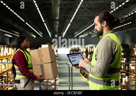 Male employee checking parcel status on a tracking software with tablet, shipment details and AWB tags ensuring package tracking in warehouse. Adult man wearing safety vest on platform. Stock Photo