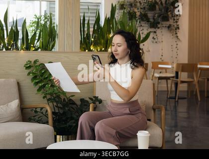 A woman sits in a modern cafe surrounded by green plants. She holds a piece of paper in one hand and looks at her phone with the other hand. Stock Photo