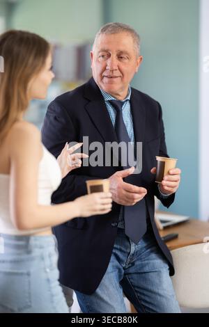 Mature boss stands in the office and talks to a young female employee, they drink coffee Stock Photo
