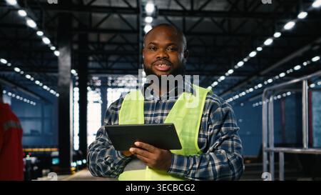 Factory supervisor checks assembly equipment on a tablet in manufacturing plant, wearing hi vis vest for the engineering industrial operation behind heavy steel product development. Stock Photo