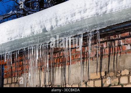 Glistening icicles cascade from a heavy snowdrift on a rooftop, contrasting sharply with the rugged brick wall and bright winter sky Stock Photo