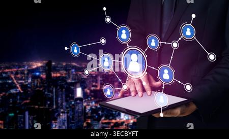 A business professional engages with a digital tablet, displaying interconnected user profiles and a vibrant city skyline at night, symbolizing techno Stock Photo
