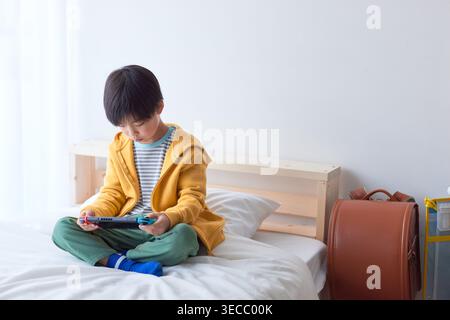 Japanese elementary school boy focused playing a portable game on a bed in a bright room Stock Photo