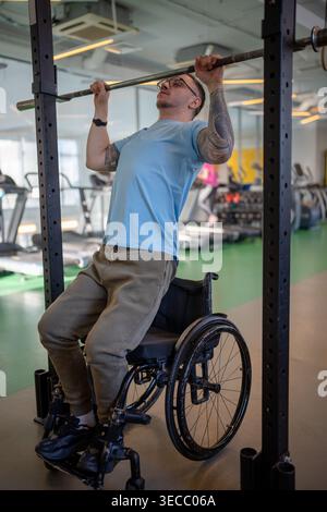 Man athlete in wheelchair doing vertical pull ups exercise using adaptive sports equipment at gym. Stock Photo