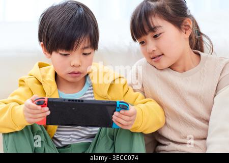 Japanese boy and girl siblings focused on portable game console indoors Stock Photo
