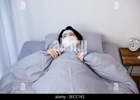 Sick young male student wearing a mask and cooling patch on his forehead in bed Stock Photo