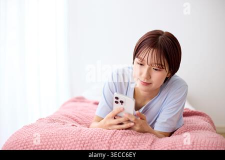 Japanese young woman operating her smartphone lying face down on bed at home Stock Photo