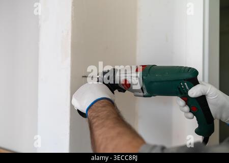 Skilled construction technician using a cordless electric drill to create holes in drywall during a home renovation. Clean and organized workspace with white walls visible in the background. Stock Photo