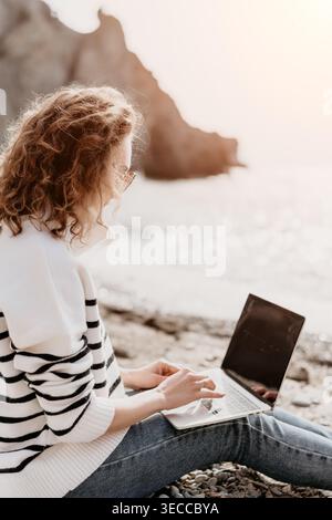 Laptop remote beach woman sits on pebbles typing on her computer with scenic ocean and cliffs in the background Stock Photo