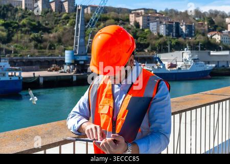 Port worker wearing safety vest checking clipboard at harbor Stock Photo