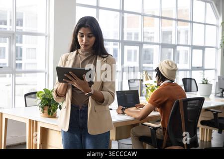 Diverse coworkers wearing beige blazer, rust shirt, working in open office using tablet laptop Stock Photo