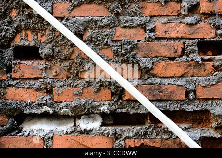 A weathered, textured red brick wall with visible mortar and missing bricks, diagonally crossed by a white pipe, showcasing an aged background Stock Photo