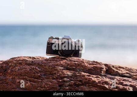 Vintage Canon AE-1 Program 35mm film camera resting on a red rock at the beach, with a blurred ocean horizon in the background Stock Photo
