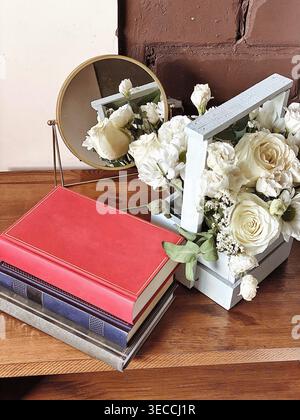 Stylish still life with a bouquet of white flowers in a wooden box, a stack of books and a round mirror on a wooden table. Soft natural light and warm Stock Photo