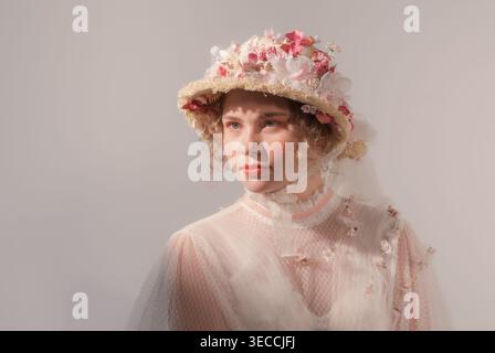Young woman models elaborate flower hat and vintage-inspired sheer top in a bright studio Stock Photo