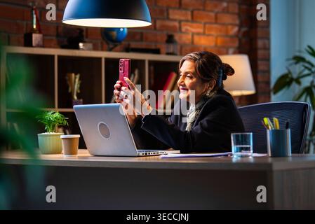 Senior Indian businesswoman in corporate attire chatting and typing on smartphone while sitting at workstation with laptop in modern cozy office space Stock Photo
