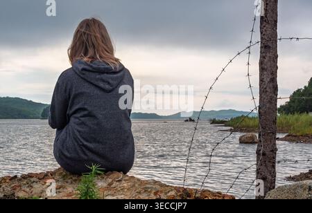 Blonde haired woman in dark hoodie sits on rugged rocks by wide lake watching distant hills under cloudy sky, with barbed wire fence nearby Stock Photo