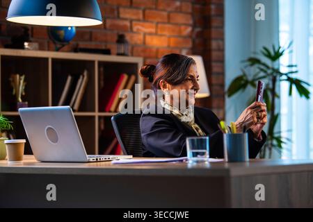 Senior Indian businesswoman in corporate attire chatting and typing on smartphone while sitting at workstation with laptop in modern cozy office space Stock Photo