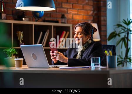 Senior Indian businesswoman in corporate attire chatting and typing on smartphone while sitting at workstation with laptop in modern cozy office space Stock Photo