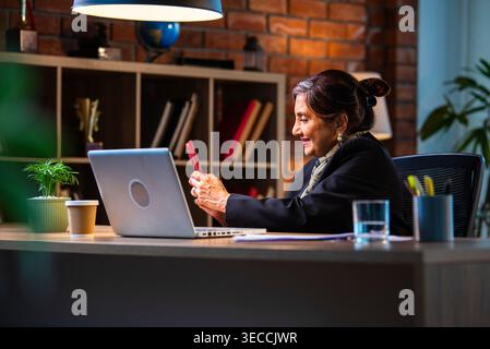Senior Indian businesswoman in corporate attire chatting and typing on smartphone while sitting at workstation with laptop in modern cozy office space Stock Photo