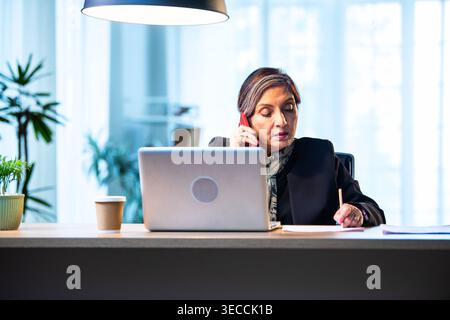 Senior Indian businesswoman in corporate attire taking notes while speaking on smartphone seated at workstation with laptop in cozy office space exper Stock Photo