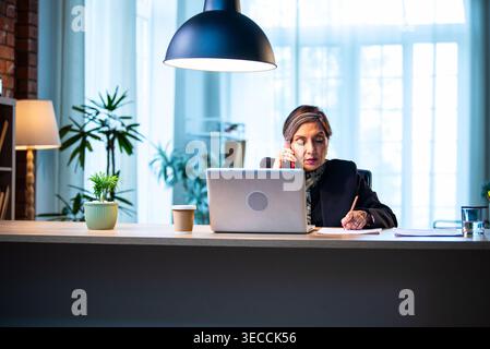 Senior Indian businesswoman in corporate attire taking notes while speaking on smartphone seated at workstation with laptop in cozy office space exper Stock Photo